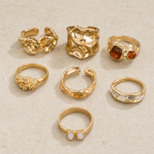 May include: A set of seven gold-toned rings featuring various designs, including organic molten metal textures, a ring with two amber-colored stones, a band with white oval inlays, and a ring with a small white stone pair.