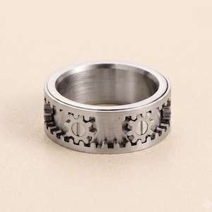 Puede incluir: Un anillo de metal plateado con un diseño de banda ancha que presenta una serie de pequeñas formas de engranajes integradas alrededor del exterior. Cada engranaje está asegurado con un detalle de tornillo central, creando un aspecto mecánico.