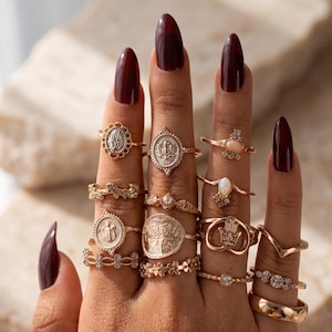 Puede incluir: Una colección de anillos dorados usados en los dedos, con varios diseños que incluyen medallones ovalados, un símbolo de mano hamsa, monedas circulares, patrones florales y pequeñas piedras claras incrustadas, creando un aspecto superpuesto.