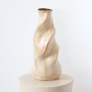 May include: A beige ceramic vase with a twisted, organic form. The vase has a narrow neck and a wider base, with a dark brown interior. It rests on a cream-coloured, round pedestal.