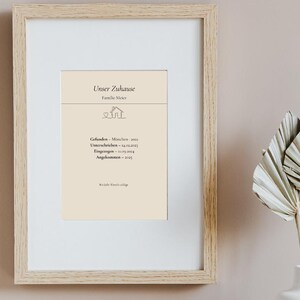 May include: A framed print with the text "Unser Zuhause" and family details. The print has a cream-coloured background and is in a light wood frame. A white vase with dried decorative leaves is to the right of the frame.
