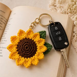 May include: A handmade crochet sunflower keychain with a gold-colored keyring and a black car key. The sunflower has yellow petals, a brown center, and green leaves. The keychain rests on an open book.