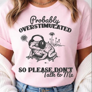 May include: Pink t-shirt with the text "Probably Overstimulated So Please Don't Talk to Me." The shirt features a black and white illustration of a frog wearing headphones. The frog is sitting down with its eyes closed.