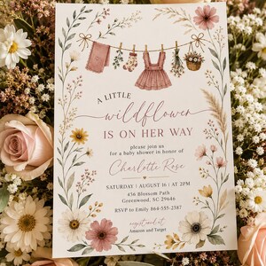May include: A rectangular baby shower invitation card featuring watercolor wildflowers and a clothesline with baby clothes. The text reads: A LITTLE wildflower IS ON HER WAY, please join us for a baby shower in honor of Charlotte Rose, SATURDAY AUGUST 16 AT 2PM, 456 Blossom Path, Greenwood, SC 29646, RSVP to Emily 864-555-2387, registered at Amazon and Target.