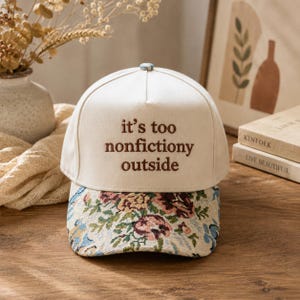 以下が含まれることがあります： クリーム色のクラウンに茶色の刺繍で「it's too nonfictiony outside」と書かれたベースボールキャップ。カーブしたつばには、青、ピンク、緑、茶色の色合いで花柄のタペストリー模様が施されています。