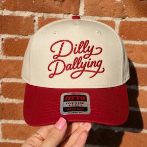 May include: A cream-colored baseball cap with a structured crown and a dark red brim. The front features the embroidered red text Dilly Dallying in a cursive script. A silver and black sticker on the brim reads Otto Five Star Premium Fabric.