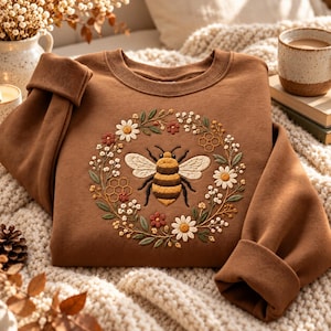 May include: A brown crewneck sweatshirt featuring a central embroidered design of a honeybee surrounded by a circular wreath of white daisies, small red flowers, green leaves, and golden honeycomb patterns stitched onto the front chest area.