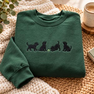 May include: A dark forest green crewneck sweatshirt featuring an embroidered design of four black Labrador puppies playing with a small light green tennis ball centered on the chest. The garment has long sleeves and a ribbed collar.