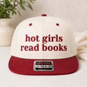 May include: A cream and burgundy baseball cap with the embroidered text "hot girls read books." The cap features a flat brim and a snap closure for an adjustable fit. The cap is made of premium fabric.
