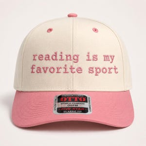 May include: A two-toned baseball cap with a cream-colored crown and a pink brim. The front features the embroidered pink text reading is my favorite sport. A silver and black sticker on the brim displays the brand name OTTO and product details.