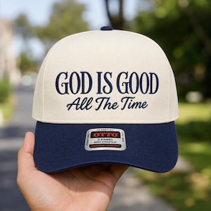 May include: A cream-colored five-panel baseball cap with a navy blue brim. The front features dark blue embroidered text that reads "GOD IS GOOD" in a bold serif font, followed by "All The Time" in a cursive script. A sticker is on the brim.
