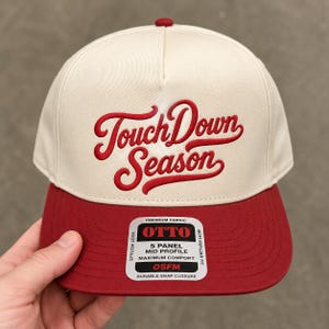 May include: A cream-colored five-panel baseball cap with a red brim and a red button on top. The front features red embroidered cursive text that reads TouchDown Season. A rectangular sticker on the brim displays OTTO branding and product details.