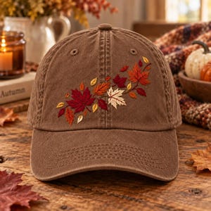 May include: A brown baseball cap featuring an embroidered design of autumn leaves in shades of red, orange, yellow, and cream. The embroidery is positioned on the front panel, showcasing detailed maple and oak leaf shapes with visible stitching.