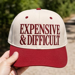 May include: A beige baseball cap with a structured crown and a solid dark red brim. The front of the cap features the embroidered text "EXPENSIVE & DIFFICULT" in a dark red serif font. The cap includes a matching dark red button on the top.