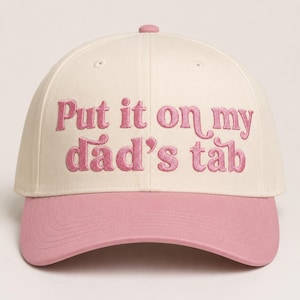 May include: Cream-colored baseball cap with a pink brim and button. The phrase "Put it on my dad's tab" is embroidered in pink script across the front. A casual, stylish accessory.