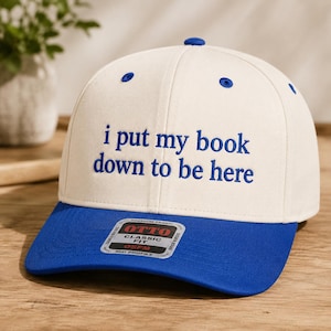 Könnte beinhalten: Eine cremefarbene Baseballkappe mit blauem Schirm und blauem Sticktext, der lautet: "i put my book down to be here". Die Kappe hat einen kleinen blauen Knopf an der Oberseite und einen rechteckigen Aufkleber auf dem Schirm mit dem Text "OTTO".