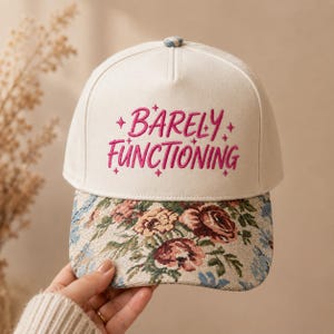 May include: A cream-colored baseball cap featuring the embroidered text "BARELY FUNCTIONING" in bright pink, accented by small star symbols. The brim displays a detailed tapestry-style floral pattern with shades of red, green, and blue.