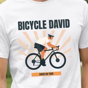 May include: White t-shirt with the text "BICYCLE DAVID" above a graphic of a cyclist on a black bicycle. The cyclist is wearing a white shirt and helmet. The text "DAVID ON TOUR" is below the bicycle graphic.