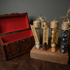 May include: A wooden treasure chest with a red felt interior sits next to a wooden block holding four glass vials. Each vial is labeled and contains dice. The labels read "Giant's Teeth", "Dragon's Blood", "Phoenix Nectar", and "Mana Juice Hour".