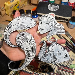 Könnte beinhalten: Vier schwarz-weiße ausgeschnittene Figuren einer Frau mit langen Haaren und einem eindringlichen Ausdruck. Die Figuren werden in einer Hand gehalten, mit Kunstbedarf und einem gemusterten Stoff im Hintergrund. Die Figuren scheinen von Manga oder Anime inspiriert zu sein.