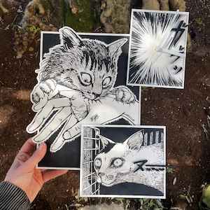 Può includere: Opere d'arte in stile manga in bianco e nero con gatti. Un'immagine mostra un gatto che morde una mano, un'altra un gatto dietro le sbarre e una terza con un quadrante e testo giapponese. L'opera è esposta contro un tronco d'albero.