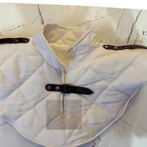 Puede incluir: Protector de cuello acolchado blanco con un patrón de diamantes y cuello alto. Cuenta con correas de cuero marrón con hebillas y fijaciones para los hombros. Un accesorio medieval.