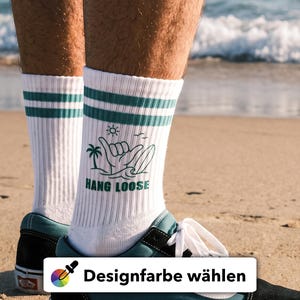 Könnte beinhalten: Weiße Socken mit zwei grünen Streifen am oberen Rand und einem grünen Grafikdruck, der eine Palme, ein Surfbrett, eine Sonne und eine Hand mit dem Shaka-Zeichen über dem Text HANG LOOSE in grünen Großbuchstaben zeigt.