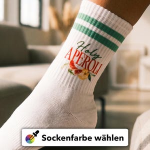 Könnte beinhalten: Weiße Socken mit grünen Streifen am oberen Rand und dem Schriftzug "Holy Aperol" in Schreibschrift, zusammen mit einer Grafik eines Aperol Spritz. Die Socken haben eine gerippte Textur und werden an einem Fuß getragen.