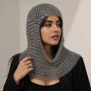 Puede incluir: Una capucha de cota de malla de color plateado construida con numerosos anillos metálicos pequeños entrelazados. La capucha cubre la cabeza y cae sobre los hombros, presentando una textura flexible similar a una malla.