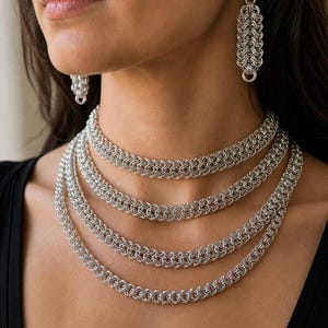 May include: A silver chainmail necklace and matching earrings. The necklace has multiple strands of interconnected rings, creating a layered effect. The earrings are a similar chainmail design, hanging from a hook.
