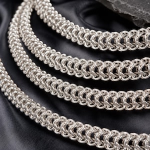 Peut inclure: Un collier en cha&icirc;ne de couleur argent&eacute;e pr&eacute;sentant un motif complexe de maillons entrelac&eacute;s. Les maillons m&eacute;talliques sont dispos&eacute;s en plusieurs rang&eacute;es parall&egrave;les, cr&eacute;ant une structure dense, textur&eacute;e et flexible avec une finition brillante.