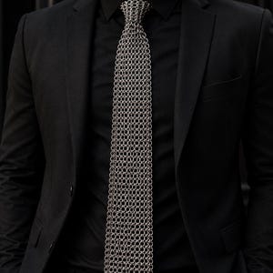 Könnte beinhalten: Eine silberne Kettenhemd-Krawatte, bestehend aus ineinandergreifenden Metallringen, hängt vor einem schwarzen Hemd und einem Anzug. Die Textur und der metallische Glanz der Krawatte bilden einen auffälligen Kontrast zur dunklen Kleidung. Die Gesamtästhetik ist modern und kantig.