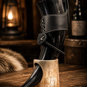 Könnte beinhalten: Ein schwarzes Trinkhorn mit Lederriemen und Halterung. Das Horn ist auf einem Holzständer montiert. Der Lederriemen ist mit weißem Faden genäht. Der Hintergrund umfasst eine Laterne und eine Flasche.