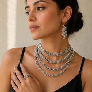 Puede incluir: Un conjunto de joyería de cota de malla plateada que consta de un collar de varias capas, un pendiente colgante a juego y un anillo. El collar presenta cuatro hileras superpuestas de anillos metálicos entrelazados.
