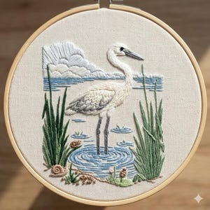 Peut inclure: Broderie artisanale représentant un héron blanc dans l'eau, entouré de roseaux verts et d'un ciel bleu nuageux. L'œuvre est encadrée dans un cercle de bois, présentant une scène de nature détaillée.