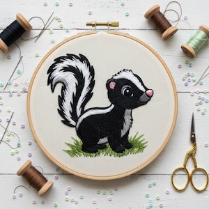Może przedstawiać: Haftowany skunks w drewnianym tamborku. Skunks jest czarno-biały z różowym nosem i uszami, stojący na zielonej trawie. Materiały do haftu, w tym szpulki nici, igły i nożyczki, otaczają tamborek na białej drewnianej powierzchni.