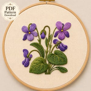 May include: A circular wooden embroidery hoop containing a cream-colored fabric with a hand-stitched design of purple violet flowers and green leaves. The text PDF Pattern Download is visible in the upper left corner of the image.