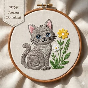 May include: A round wooden embroidery hoop containing a fabric panel with a stitched gray kitten featuring large blue eyes and pink inner ears, sitting beside a yellow and white flower stem. The text PDF Pattern Download is visible.