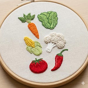 Op de afbeelding: Een borduurring met geborduurde groenten: een wortel, sla, maïs, bloemkool, een tomaat en een chilipeper. De groenten zijn geborduurd met gedetailleerde texturen en levendige kleuren, tegen een neutrale stoffen achtergrond.
