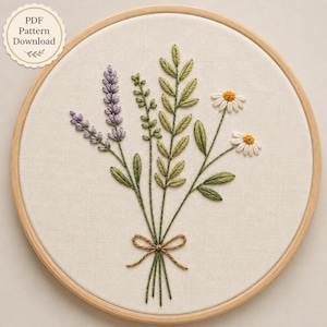 May include: A circular wooden embroidery hoop containing a fabric panel with a hand-stitched floral design. The embroidery features lavender sprigs, green leafy stems, and two white daisies with yellow centers, tied together with a brown thread bow.