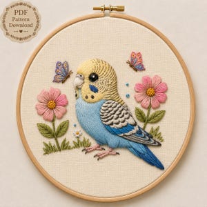 Könnte beinhalten: Ein Stickrahmen mit einem Stoffstück, das ein gesticktes Motiv eines gelb-blauen Wellensittichs, zwei rosa Blumen mit grünen Blättern und zwei kleine Schmetterlinge zeigt. Oben links befindet sich ein Abzeichen mit PDF Pattern Download.
