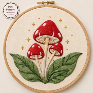 Può includere: Un telaio da ricamo contenente un pezzo di tessuto con un disegno cucito a mano di tre funghi rossi con macchie bianche, steli color marrone chiaro e grandi foglie verdi. Piccole stelle gialle ricamate circondano i funghi. Testo PDF Pattern Download.