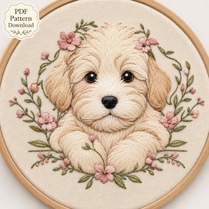 May include: A circular embroidery hoop containing a fabric panel with a stitched design of a light brown puppy surrounded by a wreath of small pink flowers and green leaves. The text PDF Pattern Download is printed in the upper left corner.