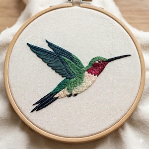 Puede incluir: Un bastidor de bordado circular de madera que contiene una tela de color blanco roto. La tela presenta un bordado detallado a mano de un colibrí en vuelo, mostrando hilos de colores verde vibrante, azul cerceta y magenta intenso.