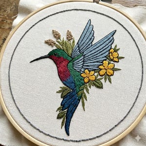 May include: A circular wooden embroidery hoop containing a fabric panel with a hand-stitched hummingbird. The bird features a red throat, green body, and blue wings. It is surrounded by green leaves, small beige buds, and three bright yellow flowers.