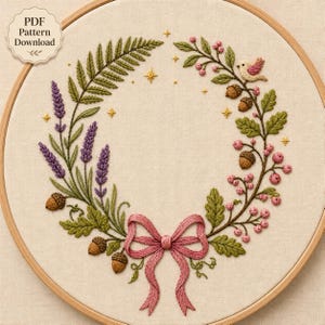 Könnte beinhalten: Ein runder Stickrahmen mit einem gestickten Kranzmotiv auf beigem Stoff. Der Kranz besteht aus violettem Lavendel, grünen Farnblättern, braunen Eicheln, rosa Beeren, einem kleinen Vogel und einer rosa Schleife, verziert mit kleinen goldenen Sternen.