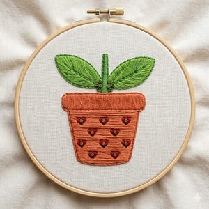 Pode incluir: Design bordado de vaso de plantas em tecido branco, num bastidor de bordado de madeira. O vaso é laranja com detalhes vermelhos em forma de coração. Folhas e caule verdes erguem-se do vaso, criando uma estética charmosa e artesanal.