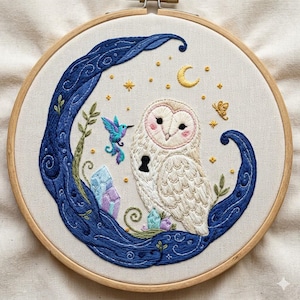 Könnte beinhalten: Ein runder Stickrahmen mit einem weißen Stoffeinsatz, der ein handgesticktes Motiv einer weißen Eule mit einem schwarzen Schlüsselloch auf der Brust, einem blauen Kolibri, violetten und blauen Kristallen sowie einem gelben Halbmond zeigt.