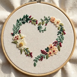 May include: A circular wooden embroidery hoop containing off-white fabric with a hand-stitched heart-shaped wreath. The design features green leaves, small red berries, and various flowers in shades of cream, yellow, and soft peach.