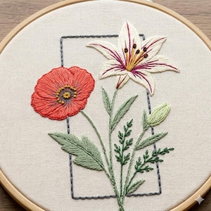 May include: An embroidery hoop containing a piece of off-white fabric with a hand-stitched design. The design features a bright red poppy flower and a white lily with purple accents, surrounded by green stems and leaves inside a dark gray rectangle.